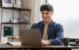 Happy Arabic Man Using Laptop Typing And Browsing Internet Sitting At Table In Office. Young Businessman Working Online On Computer At Modern Workplace. Entrepreneurship Concept photo