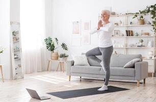 Sporty Senior Woman Practicing Yoga With Online Tutorials At Home, Standing In Tree Pose And Smiling, Enjoying Retirement Time, Free Space photo