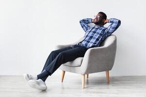 Peaceful young black man relaxing in armchair against white studio wall, free space. Full length of African American guy enjoying lazy weekend morning, resting with hands behind head photo