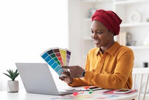 Stylish young black woman in red turban independent contractor having conference with client, female designer sitting at workdesk in front of laptop at home, showing color palette, copy space photo