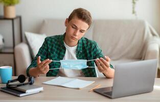 Pensive bored teen guy holding protective mask, studying at home during lockdown, empty space photo