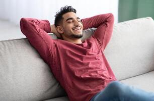 Happy middle-eastern guy in casual chilling at home, reclining on couch with closed eyes and smiling. Relaxed young arab man enjoying his new comfortable couch, leaning back, copy space photo