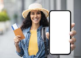 Happy attractive millennial brunette lady in straw hat tourist holding passport with flight tickets and cell phone with white blank screen, showing nice mobile app for travellers, mockup photo