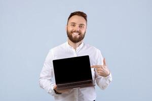 Happy young businessman pointing at laptop with mockup for website design on screen, blue studio background. Joyful male entrepreneur showing computer with space for your advertisement photo