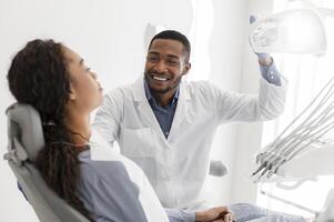 Cheerful black dentist turning on equipment before check up and greeting female patient photo