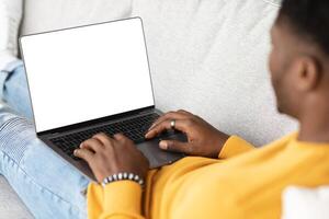 Unrecognizable black guy using modern laptop with empty screen, typing on computer keyboard, reclining on couch, using nice software, programming, chatting with ladies, cropped, mockup photo