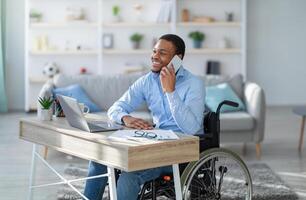Joyful disabled black guy in wheelchair having phone conversation while working online on laptop from home. Young handicapped businessman talking with client on mobile phone photo