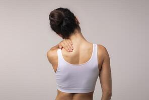 Unrecognizable brunette woman with neck and back pain rubbing her painful body neck area, lady suffering from office syndrome, back view, grey studio background, copy space photo