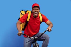 Fast Delivery Service. Overjoyed african american courier guy in red cap and uniform riding bicycle and wearing thermo backpack bag isolated on blue studio background. Shipment service during lockdown photo