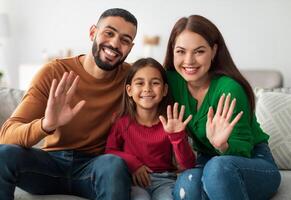 Hello. Cheerful Young Middle Eastern Family Of Three People Waving Hand Smiling To Camera Sitting On Couch At Home. Hi Greeting Gesture Concept. Call And Modern Remote Communication photo