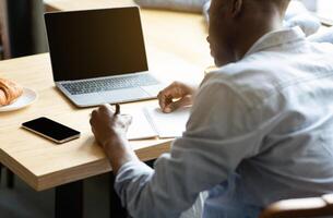 Unrecognizable black guy using laptop with blank screen, studying online, taking notes in copybook at cafe, mockup for design. Millennial man working remotely at coffee shop, space for website photo