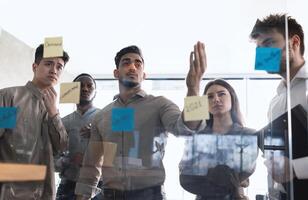 enfocado diverso grupo de negocio personas que se discute idea o tarea, mirando a enviar eso pegajoso notas en vaso pared. grave equipo desarrollando trabajo plan en creativo corporativo oficina a estar arriba reunión foto