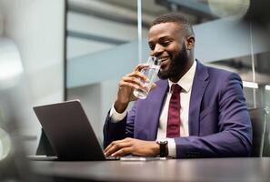 Positive young black man manager drinking water while working on laptop, typing on computer keyboard, making marketing research or sending emails, office interior, copy space photo