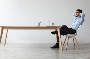 Taking Break. Profile side view of smiling young Arab man relaxing on chair sitting at table and using laptop, happy millennial male leaning back at workplace, looking at pc screen watching photo