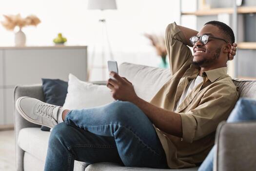 nuevo móvil aplicación alegre negro hombre utilizando Teléfono móvil vistiendo los anteojos, mensajes de texto y hojeada Internet sentado en sofá a hogar. teléfono usuario utilizando solicitud adentro. lado ver foto