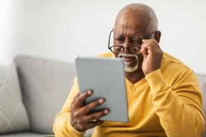 Senior Black Man With Poor Eyesight Using Digital Tablet Wearing Glasses And Squinting Eyes Reading News Online Sitting On Couch At Home. Eyes Health Issues Concept. Selective Focus photo