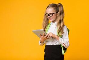 Gadgets And School. Cheerful Schoolgirl Using Tablet Computer Browsing Internet Over Yellow Background. Copy Space photo