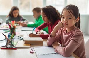 Unhappy Asian Schoolgirl Thinking Having Problem With Education Sitting While Her Diverse Classmates Learning Lesson In Modern Classroom At School. Educational Issues Concept. Selective Focus photo