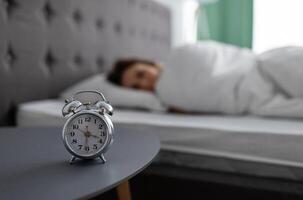 Alarm clock on bedside table and blurred view of millennial lady lying in bed, covered with warm blanket, unwilling to wake up, selective focus. Young woman feeling sleepy, free space photo