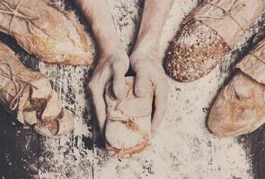 Lots of different bread sorts, wrapped in craft paper. Baking and cooking concept background. Hands of baker carefully hold loaf on wooden table, sprinkled with flour. photo
