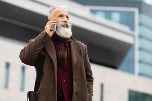 Wealthy senior grey-haired man with long beard having conversation on mobile phone while walking by street, standing by business center, going to business meeting, looking at empty space photo