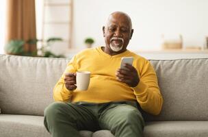 Mature African American Male Using Cellphone Browsing Internet And Texting Communicating Online In Social Media App Sitting On Sofa At Home. Senior Man Drinking Coffee And Scrolling News On Phone photo