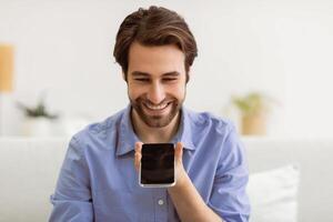 Voice Assistant App. Cheerful Man Using Smartphone With Voice Search Application Sitting On Couch At Home. Guy Chatting On Cellphone Using Loudspeaker Mode. Modern Gadgets Lifestyle Concept. photo