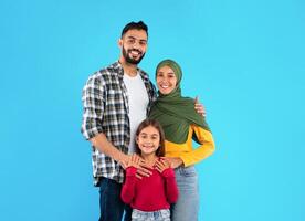 Modern Muslim Family Of Three Hugging Smiling To Camera Standing Together Over Blue Studio Background. Islamic Mother In Hijab And Father Embracing Their Happy Little Daughter photo