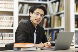 Working On Research Project. Portrait of asian male student using laptop at library, writing in notepad, looking at camera photo