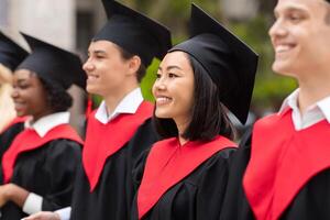 Multiracial group of cheerfully graduates having graduation ceremony, wearing black and red graduation robes and caps, standing in a row at university campus, closeup, panorama photo