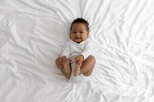 Top View Of Cute Little Black Baby Lying On Bed At Home, Adorable African American Infant Boy Having Fun In Bedroom, Playing With His Toes And Looking At Camera While Resting On White Bedsheet photo