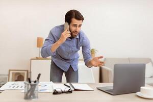 Displeased Businessman Talking On Cellphone Having Computer Problem Standing In Modern Office. Discontented Customer, Communication Issue, Problems At Work Concept. Selective Focus photo