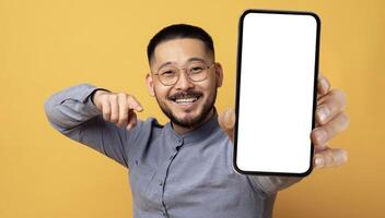 Mobile Ad. Smiling Young Asian Man Pointing At Big Blank Smartphone In His Hand While Standing Over Yellow Studio Background, Happy Millennial Guy Recommending New App Or Website, Collage, Mockup photo