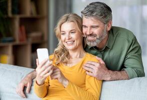 Portrait Of Smiling Middle Aged Couple Resting With Smartphone On Couch, Happy Mature Spouses Watching Photos Or Browsing New Mobile App While Relaxing In Living Room, Closeup Shot With Free Space