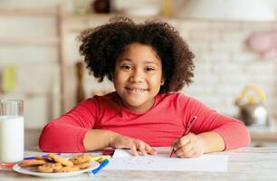 Portrait Of Happy Cute Little Black Girl Drawing In Kitchen, Adorable African American Female Child Sitting At Table With Cookies And Milk And Smiling At Camera, Closeup Shot With Free Space photo