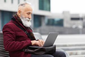 Senior bearded businessman with modern laptop sitting on bench, typing on keyboard, sending email, empty space. Stylish aged grey-haired man sitting on the street by business center, working online photo