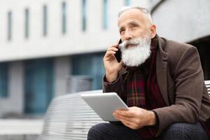 Elderly grey-haired man with long beard sitting on bench by office building, holding digital tablet and talking on mobile phone, having business call while resting on the street, looking at free space photo