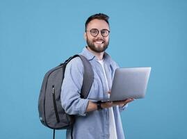 Smart millennial student with backpack using laptop computer over blue studio background. Cheerful young guy doing online work, communicating with family or friend, attending webinar photo
