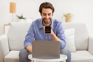 Cheerful Guy Using Voice Assistant On Mobile Phone Or Talking Using Loudspeaker Mode On Smartphone Sitting At Laptop Computer On Sofa At Home. Voice Search Application, Gadgets Lifestyle. photo