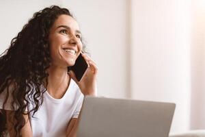 Closeup of happy pretty lady talking on mobile phone and looking at copy space, using laptop. Joyful curly woman working from home, having conversation with colleagues or clients on phone photo
