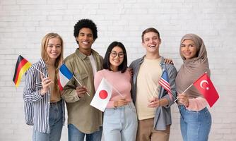 Student exchange, international friendship. Multiethnic young students with flags of different countries ready to study and look at camera on brick white wall background, studio shot, free space photo