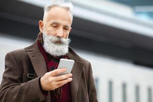 Grey-haired elderly businessman using smartphone on the street, checking his agenda, copy space. Bearded aged man using mobile phone, websurfing or chatting with colleagues, having lunch break photo
