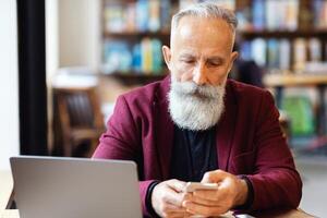 Busy aged grey-haired businessman with smartphone sitting in front of modern laptop at cafe, working while having coffee break, empty space. Successful senior man in suit checking email photo