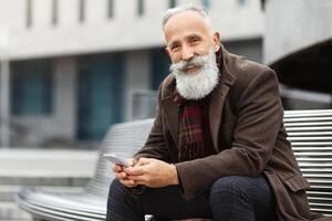 Smiling senior grey-haired man with mobile phone sitting on bench on street, smiling at camera, copy space. Good-looking aged man with long beard resting by office building, having lunch break at job photo