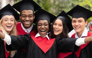 Happy multiracial group of students in graduation dresses and hats taking selfie together, posing at university campus, enjoying and celebrating graduation, closeup portrait photo