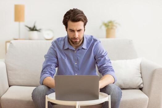 Man Frowning Using Laptop Computer Having Problem Browsing Internet Sitting On Sofa At Home. Unemployed Guy Searching Employment Online, Internet Connection Issue Concept photo