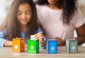 Eco-friendly family activities. Lovely little black girl with her mother playing garbage sorting board game at home, selective focus. Adorable child putting toy trash into tiny bins photo