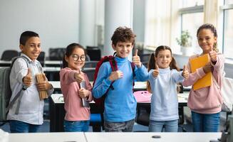 Education Concept. Diverse group of happy smiling international classmates standing in line with notebooks and backpacks in classroom and showing thumbs up sign gesture, enjoying studying at school photo