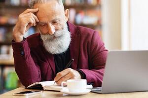 Thoughtful aged businessman with grey hair checking agenda, taking notes in notebook, working with laptop at cafe, copy space. Elderly concentrated man looking for job, checking his list, drinking tea photo