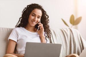 Closeup of smiling young woman freelancer talking on mobile phone and using laptop, working from home during isolation, looking at copy space. Happy curly lady having conversation on phone photo